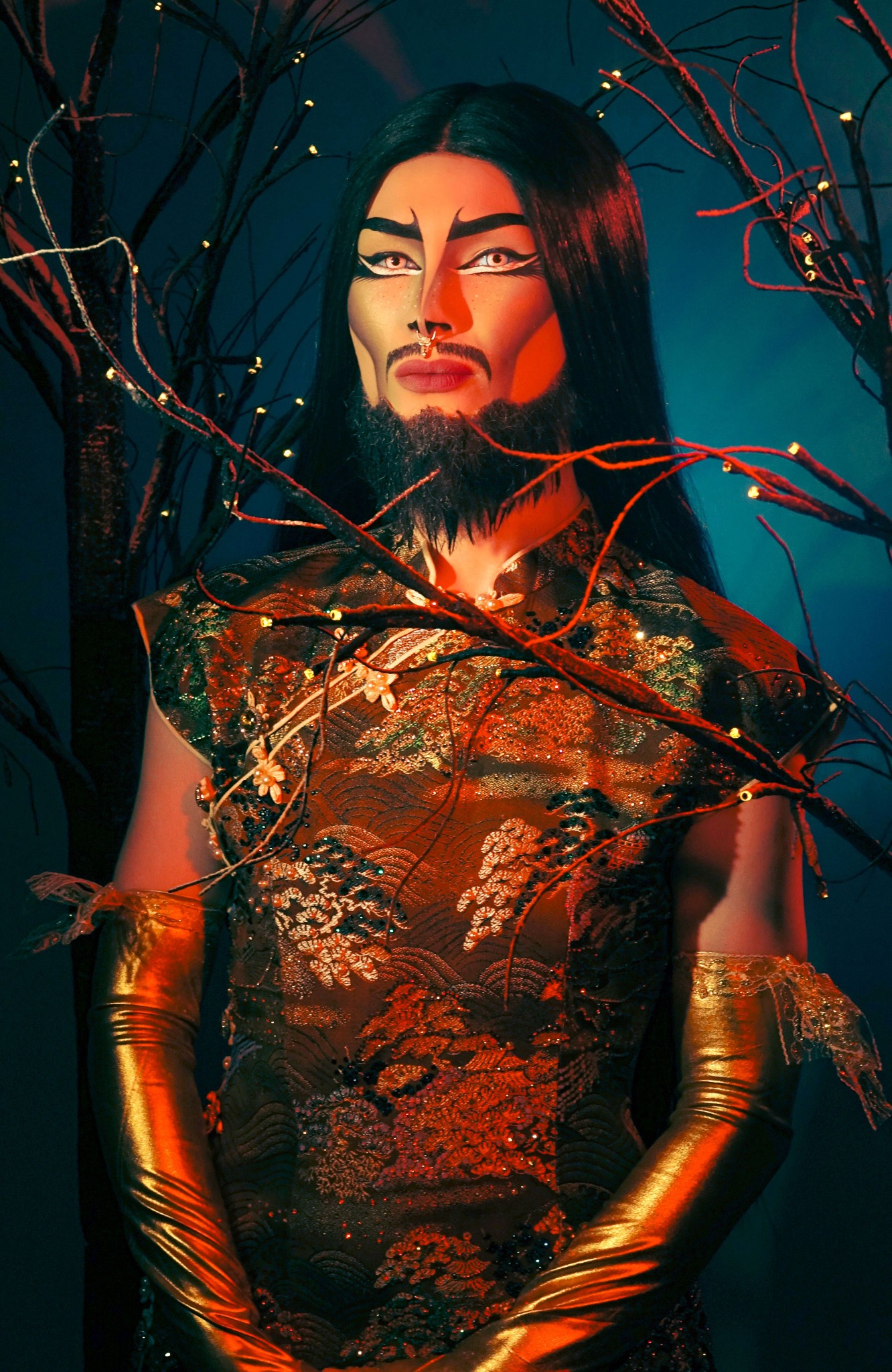 KAANYAG, 2025 portrait of a drag performer with black hair, beard, gold gloves, and branches